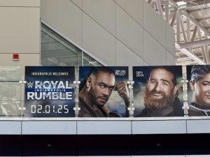 Several graphics of WWE Superstars hang above on a bridge at the airport. This photo shows the "WWE Royal Rumble 2025" logo, Damian Priest, Sami Zayn, and Bayley.