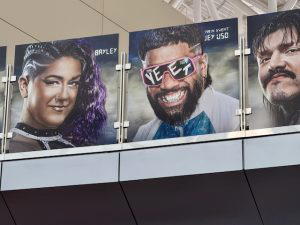 Several graphics of WWE Superstars hang above on a bridge at the airport. This photo shows Bayley, Jey Uso, and Dominik Mysterio.