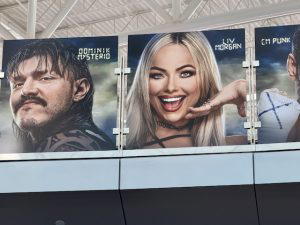 Several graphics of WWE Superstars hang above on a bridge at the airport. This photo shows Dominik Mysterio, Liv Morgan, and CM Punk.