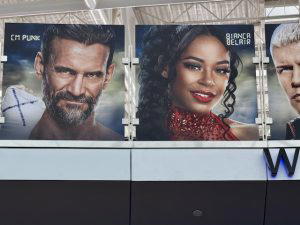 Several graphics of WWE Superstars hang above on a bridge at the airport. This photo shows CM Punk, Bianca Belair, and Cody Rhodes.