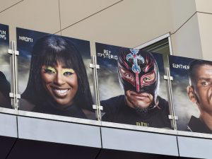 Several graphics of WWE Superstars hang above on a bridge at the airport. This photo shows Naomi, Rey Mysterio, and Gunther.